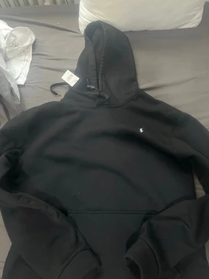 Svart hoodie från Polo Ralph Lauren - Snygg svart hoodie från Polo Ralph Lauren med klassisk huva och dragsko. Liten broderad logga på bröstet i vitt. Tillverkad i mjukt material som känns skönt mot huden. Perfekt för en avslappnad och stilren look.