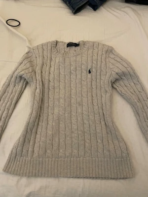 Beige kabelstickad tröja Polo Ralph Lauren - Kabelstickad tröja i beige från Polo Ralph Lauren. Klassisk modell med rund halsringning, ribbade muddar och det ikoniska mörkblå logobroderiet på bröstet. Tillverkad i mjukt material som ger en skön och stilren look.