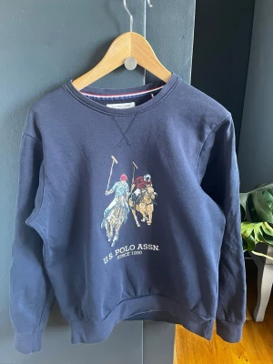 Svart sweatshirt U.S. Polo Assn S - Svart sweatshirt från U.S. Polo Assn. Med stort tryck, Rund halsringning och långa ärmar. Klassisk och stilren design, perfekt för dig som gillar detaljer.