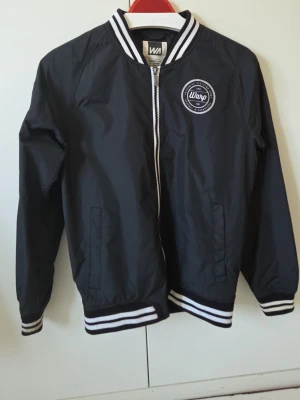 Svart bomberjacka från Warp - Snygg svart bomberjacka från Warp med vita ränder på krage, ärmslut och nederkant. Jackan har dragkedja framtill, två sidofickor och en rund logga på bröstet. Tillverkad i ett lätt och slitstarkt material, perfekt för en cool streetstil.