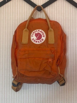 Orange Kånken ryggsäck Fjällräven - Säljer en klassisk orange Kånken ryggsäck från Fjällräven. Väskan har det ikoniska runda logotypmärket framtill, justerbara beige axelband och bärhandtag. Tillverkad i slitstarkt syntetmaterial med en stor ficka fram och dragkedja.