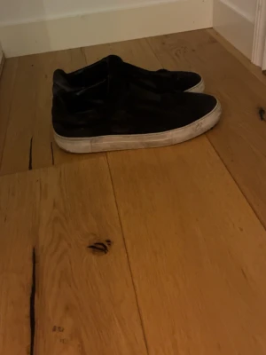 Svarta sneakers från Axel Arigato - Säljer ett par svarta sneakers från Axel Arigato med vit sula och slip-on design. Skorna har en stilren look med ovandel i mocka och elastiska band över vristen för enkel påtagning. Perfekta för dig som gillar minimalistisk och modern stil. Pris kan diskuteras 