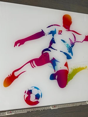 Färgglad tavla med fotbollsmotiv - Säljer en modern tavla med en färgstark illustration av en fotbollsspelare som sparkar en boll. Tavlan har vita bakgrund och motivet går i rött, blått, orange och grönt. Perfekt för dig som gillar sportig inredning eller vill ha något unikt på väggen.