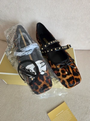 Leopardmönstrade ballerinaskor med nitar - Snygga ballerinaskor från Michael Kors i leopardmönstrad kalvhårsläder. Skorna har en fyrkantig tå, dekorativ rosett framtill och ett Mary Jane-band med guldfärgade nitar. Perfekt för dig som vill sticka ut med en trendig och djurinspirerad look.