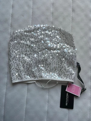 Vit paljettprydd tubtopp PrettyLittleThing - Säljer en vit tubtopp från PrettyLittleThing täckt med glittriga silverpaljetter. Toppen är axelbandslös och har en bandeauformad passning med elastisk kant för att sitta på plats. Perfekt för dig som vill sticka ut och glänsa.