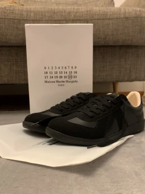 Maison Margiela svarta sneakers - Svarta sneakers från Maison Martin Margiela med en clean och stilren design. Skorna har en mix av mocka och läderdetaljer, svarta skosnören och en låg siluett. Perfekta för dig som gillar minimalistisk och exklusiv streetwear. Boxen e lite sönder!
