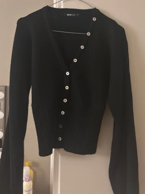 Svart kofta från Gina Tricot - Snygg svart kofta från Gina Tricot med v-ringning och silvriga knappar hela vägen framtill. Modellen är croppad med bred ribbstickad midja och långa ärmar. Perfekt att slänga över en topp för en stilren look.