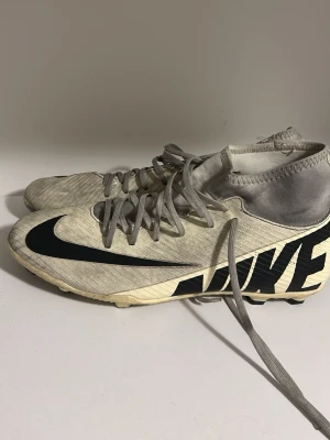 Nike vita fotbollsskor med högt skaft - Säljer ett par vita Nike fotbollsskor med högt skaft och stora svarta loggor på sidan och tån. Skorna har snörning och är tillverkade i syntetmaterial för bra grepp och passform på planen. Perfekta för dig som vill ha stilrena och funktionella skor till matchen.