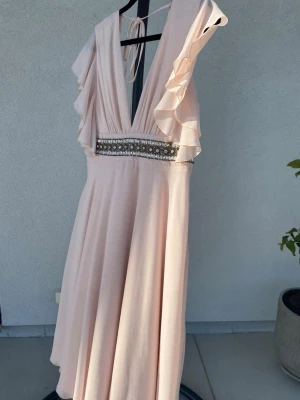 Ljusrosa klänning  med volang - Elegant ljusrosa tre kvarts längd  med djup v-ringning och volangdetaljer längs axlarna. Klänningen har ett brett midjeband dekorerat med pärlor och paljetter samt öppen rygg med knytband. Perfekt för speciella tillfällen. Uk size 12