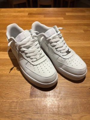 Nike Air Force 1 vita sneakers - Klassiska Nike Air Force 1 sneakers i helvitt läder med perforerad tåbox, tjock sula och ikonisk Swoosh på sidan. Skorna har vita skosnören och en stilren, tidlös design som passar till det mesta. Perfekta för dig som gillar streetwear och sneakers.