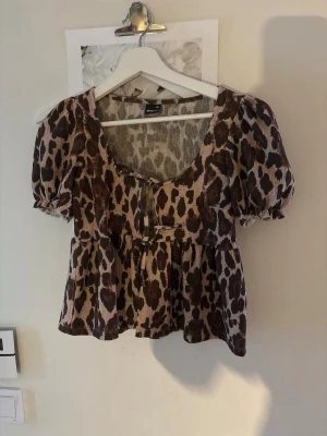 Leopardmönstrad blus med puffärm - Säljer en leopardmönstrad blus med puffiga korta ärmar och söt knytning framtill. Blusen har en v-ringad urringning och volang nedtill för en trendig look. Perfekt för dig som vill sticka ut lite extra.