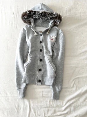Grå hoodie med fuskpäls från Hollister - Snygg grå hoodie från Hollister med knappar framtill och broderad logga på bröstet. Huvan har fuskpäls och dragsko, samt ribbade muddar vid ärmslut och nederkant. Perfekt för dig som gillar en casual och mysig stil.