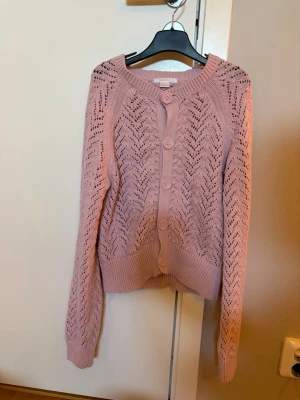 Rosa kofta från Gina Tricot - Söt rosa kofta från Gina Tricot med stora knappar framtill och ett snyggt hålmönster. Koftan har ribbade muddar vid ärmslut och nederkant samt rund halsringning. Perfekt för lager på lager och ger en trendig touch till din outfit.