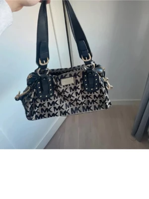 Beige och svart MK handväska - Snygg handväska från Michael Kors i beige och svart med tydligt MK-mönster över hela väskan. Väskan har svarta detaljer, guldfärgade metalldelar och dubbla axelband. Materialet är canvas med läderdetaljer och nitar på handtagen för en cool look. Köpt för 900kr och ser ut likadan ut sen jag köpte den
