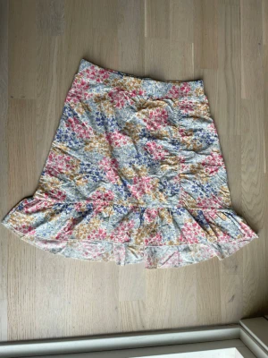 Blommig volangkjol i bomull - Söt kortkjol med volangkant och blommigt mönster i rosa, blått och gult på vit botten. Kjolen är gjord i mjuk bomull och har en avslappnad passform som ger en luftig känsla. Perfekt för dig som gillar färgglada och romantiska plagg.