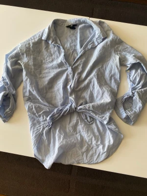 Ljusblå knytblus från H&M - Säljer en ljusblå skjorta från H&M med knyt framtill. Skjortan har lång ärm med knapp vid ärmslut och klassisk krage. Perfekt till jeans eller kjol för en avslappnad look.