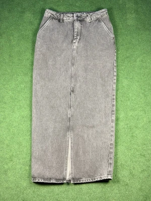 Grå lång jeanskjol med slits - Säljer en grå lång jeanskjol med hög midja och slits framtill. Kjolen har klassiska fickor och bälteshällor, perfekt för en avslappnad och trendig look. Materialet är kraftigt denim som ger en snygg vintagekänsla.