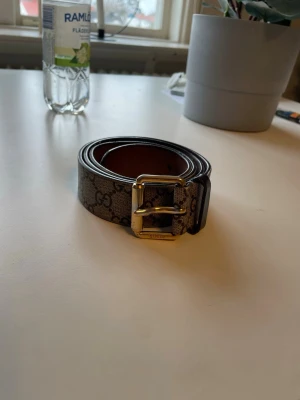 Gucci bälte med GG-mönster och guldspänne - Snyggt Gucci bälte med klassiskt GG-mönster i beige och brunt. Bältet har ett stilrent guldigt spänne med Gucci-logga. Perfekt accessoar för att lyfta din outfit och ge en exklusiv touch.