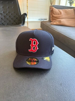 Mörkblå Boston Red Sox keps New Era - Säljer en mörkblå 59FIFTY keps från New Era med Boston Red Sox logga i rött och vitt framtill. Lätt böjd skärm och broderade detaljer. Kepsen är i ett robust material och har officiella MLB-märken på insidan.