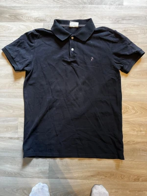 Svart pikétröja med krage - Klassisk svart pikétröja med krage och två knappar framtill. Tröjan har korta ärmar och en liten broderad logga på bröstet. Perfekt till en avslappnad men ändå stilren look.