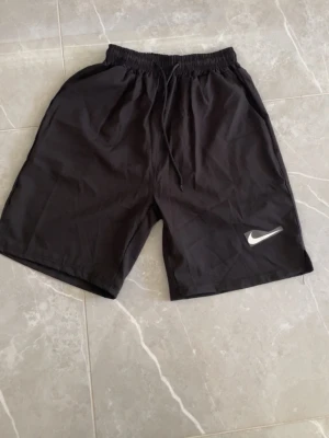 Svarta Nike shorts med snörning - Säljer ett par svarta shorts från Nike med klassisk vit logga på benet. Shortsen har elastisk midja med snörning för skön passform och är tillverkade i ett lätt syntetmaterial. Perfekta för träning eller chill dagar.