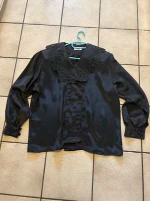 Svart blus med spetskrage storlek XL - Stilren svart blus i glansigt material med stor dekorativ spetskrage och knappar framtill. Blusen har långa ärmar med breda manschetter och en lös passform. Perfekt för dig som vill ha en trendig och unik look.