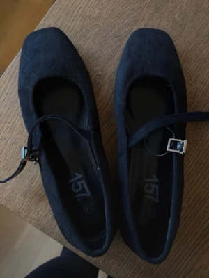 Svarta ballerinaskor med rem från 157 - Stilrena svarta ballerinaskor från 157 med fyrkantig tå och justerbar rem med silverfärgat spänne. Skorna har en mjuk yta som liknar mocka och en klassisk design som passar till många olika outfits.