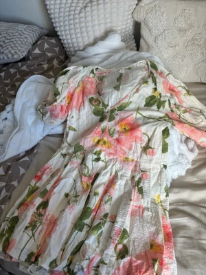 Blommig kortklänning med v-ringning - Superfin kortklänning i vitt med stora blommor i rosa, gul och grönt. Klänningen har v-ringning, trekvartsärm med resår och ett luftigt, lätt material som känns perfekt för sommaren. Passformen är normal och kjolen har ett fint fall.