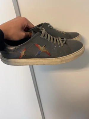 Grå sneakers med broderade fåglar - Snygga grå sneakers från Axel Arigato med broderade färgglada fåglar på sidan. Skorna har vita sulor och klassisk låg design med snörning. Tillverkade i läder för en stilren look.