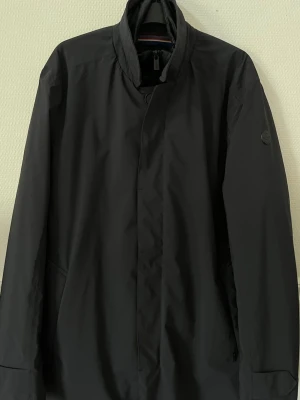 Svart jacka från snoot - Nypris 2900kr. Stilren svart jacka från snoot med hög krage och dold dragkedja samt knäppning. Rocken har en innerjacka med quiltad struktur och en orange detalj på innerfickan. Perfekt för kyliga dagar och ger en clean look.