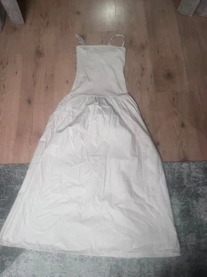 Lång vit klänning med smala band - Säljer en enkel och stilren långklänning i vitt med smala axelband och rak överdel. Klänningen har en lätt utställd kjol och är perfekt för varma dagar. Materialet känns mjukt och luftigt, troligen bomull eller linne.