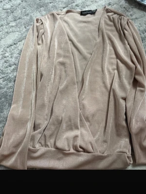 Beige omlottopp från Boohoo - Snygg beige omlottopp från Boohoo i glansigt, mjukt material. Toppen har långa ärmar med lätt puff vid axlarna och en djup v-ringning. Perfekt att styla med jeans eller kjol för en trendig look.
