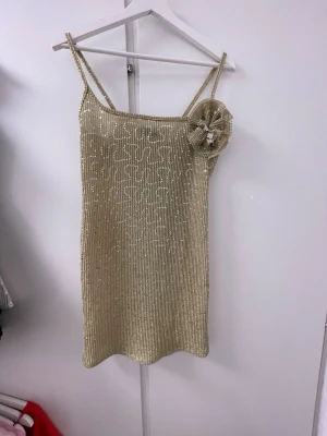 Guldig paljettklänning från ASOS DESIGN - Kort guldig klänning från ASOS DESIGN med smala axelband och dekorativ blomdetalj på axeln. Klänningen är täckt av paljetter som ger en glittrig look och har en figurnära passform. Passar perfekt för en utgång
