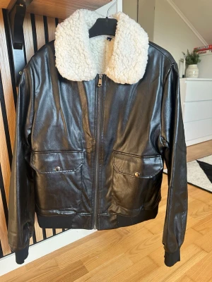 Svart skinnjacka med fuskpäls-krage - Snygg svart skinnjacka med vit, fluffig fuskpäls-krage. Jackan har två stora fickor framtill med lock och knapp, dragkedja och ribbade muddar vid ärmslut och midja. Perfekt för dig som vill ha en klassisk men trendig look.