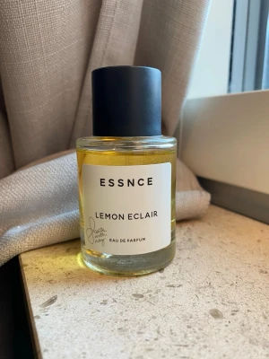 ESSNCE Lemon Eclair Eau de Parfum - Fräsch parfym från ESSNCE med doften Lemon Eclair. Flaskan är rund och genomskinlig med svart lock och vit etikett. Innehållet har en gul ton och designen är stilren och modern. Perfekt för dig som gillar citrusdofter.