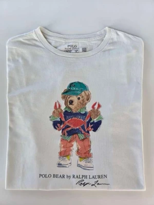 Vit Polo Bear t-shirt från Ralph Lauren - Vit t-shirt från Ralph Lauren med ikoniskt Polo Bear-tryck framtill. Björnen har på sig färgglada kläder och håller en krabba. T-shirten har rund hals och klassisk passform. Perfekt för dig som gillar streetstyle och vill sticka ut. 100 % bomull  med QR-kod