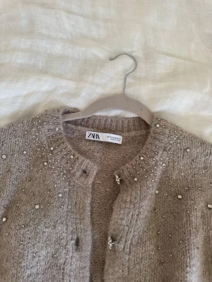 Beige kofta med pärlor från Zara - Supersöt beige kofta från Zara med glittrande pärlor och stenar på axlar och runt halsen. Koftan har rund hals, långa ärmar och fina knappar framtill. Mjuk stickad kvalitet som känns lyxig och passar perfekt till lager på lager. Passar allt från xs-m. Helt oanvänd och säljs inte längre 💗