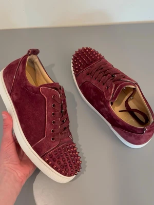 Vinröda sneakers med nitar - Säljer ett par vinröda sneakers från Christian Louboutin i mocka med coola nitar på tån. Skorna har vita sulor, matchande vinröda skosnören och en låg profil. Perfekta för dig som vill sticka ut med en edgy och lyxig stil.
