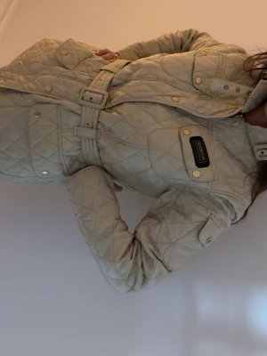 Beige quiltad jacka från Barbour - Snygg beige quiltad jacka från Barbour med bälte i midjan och guldiga knappar. Jackan har flera fickor, hög krage och Barbour-logga på bröstet. Perfekt för dig som vill ha en klassisk och stilren look.
