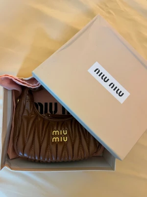Brun handväska från Miu Miu - Snygg brun handväska från Miu Miu i quiltat läder med guldiga detaljer och logotyp framtill. Kommer med original dustbag och box. Väskan har en rundad form och kort handtag, perfekt för att bära i handen eller på armen.