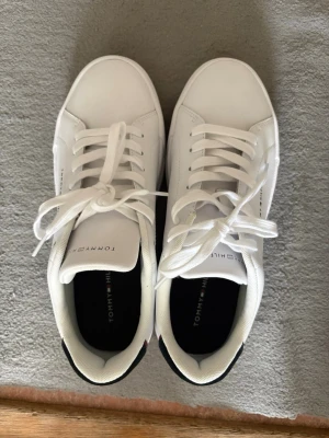 Vita Tommy Hilfiger sneakers - Fräscha vita sneakers från Tommy Hilfiger med klassisk låg modell. Skorna har vita snören, diskret logga på plösen och en snygg detalj i mörkblått och rött på hälen. Tillverkade i slätt syntetmaterial med bekväm innersula och stilren design.