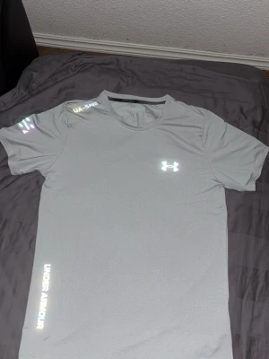 Grå Under Armour t-shirt - Snygg vit t-shirt från Under Armour med tryck på bröstet och längs sidan. T-shirten har rund hals, korta ärmar och är tillverkad i ett lätt och funktionellt material som passar perfekt till träning.