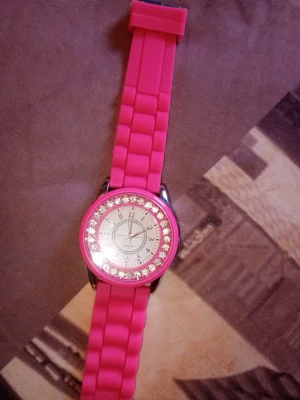 Rosa klocka med glittriga stenar - Cool rosa klocka med silikonarmband och glittriga stenar runt urtavlan. Urtavlan är vit med silverdetaljer och tydliga siffror. Perfekt accessoar för att sticka ut och ge din outfit lite extra bling.
