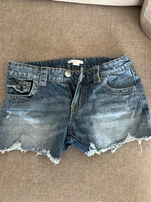 Blå jeansshorts  - Snygga blå jeansshorts med fransig, rå kant och slitningar. Klassisk femficksmodell med knapp och dragkedja framtill samt fickor med lock och knapp bak. Perfekta för sommaren och ger en avslappnad vibe.