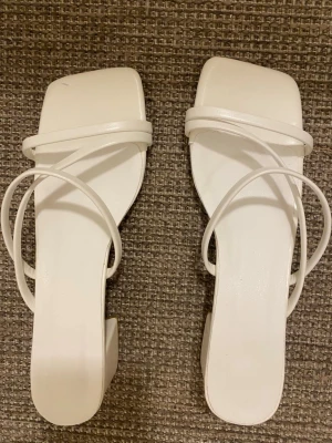 Vita sandaler med blockklack - Säljer ett par trendiga vita sandaler med fyrkantig tå och smala remmar. Sandalerna har en stabil blockklack och minimalistisk design. Perfekta till sommarens outfits och lätta att matcha. Materialet är troligtvis konstläder. Har bara använt en gång till skolavslutningen.