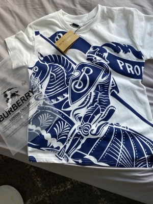 Vit Burberry t-shirt med blått tryck - Snygg vit t-shirt från Burberry med stort blått tryck framtill som föreställer en riddare och sköld. Klassisk rund halsringning och korta ärmar. Perfekt för dig som vill sticka ut med en exklusiv och trendig look.