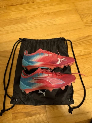 PUMA ULTRA 6 ULTIMATE FG RE-CHARGE - BLÅ/SUNSET PINK/GARNET ROSE - Säljer ett par snygga Puma Ultra fotbollsskor i rosa och blått med vita detaljer. Skorna har låg profil, snörning och är tillverkade i lätt syntetmaterial för maxad fart på planen. Perfekta för dig som vill sticka ut och spela snabbt. Påse är inkluderande med köpet!