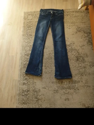 Blå bootcut jeans med brodyr - Säljer ett par blå bootcut jeans med broderade bokstäver på bakfickorna. Jeansen har klassisk femficksdesign, låg midja och utsvängda ben. Perfekta för dig som gillar en retrostil och vill sticka ut lite extra.