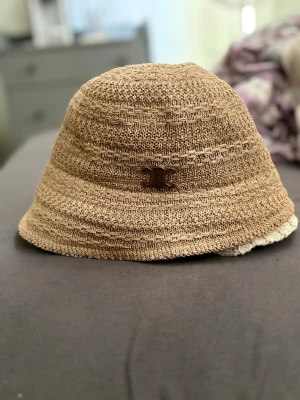 Beige stråhatt C  - Snygg beige stråhatt från C med det ikoniska Triomphe-emblemet i metall framtill. Hatten har en klassisk bucket-modell och är tillverkad i flätad strå, perfekt för soliga dagar och trendiga outfits.