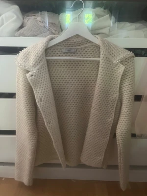 Krämvit stickad cardigan med pärlor - Superfin krämvit stickad cardigan från Mango med pärldetaljer på kragen. Cardiganen har en öppen front, grovstickad struktur och lång ärm. Perfekt för lager-på-lager och ger en lyxig touch till din outfit.